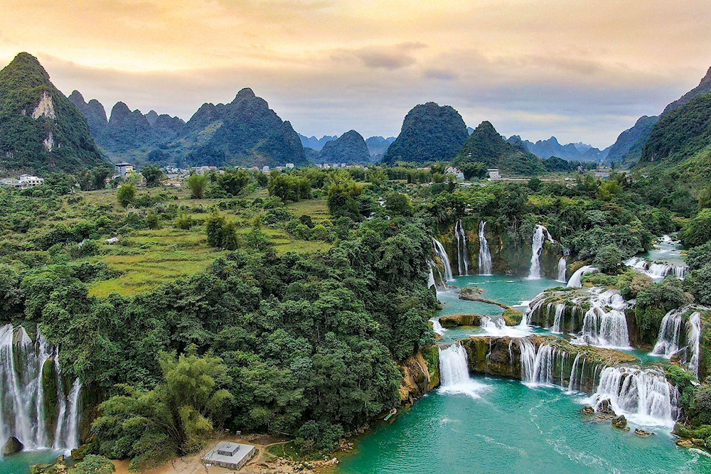 The main waterfall cascades down uneven limestone steps, creating soft white cascades that flow between lush green hills and rocky outcrops